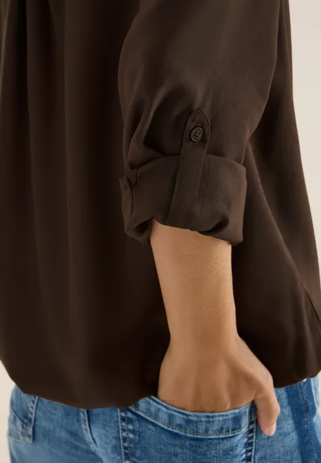 Braune Bluse mit hochgekrempeltem Ärmel, Hand steckt in blauer Jeans-Tasche.